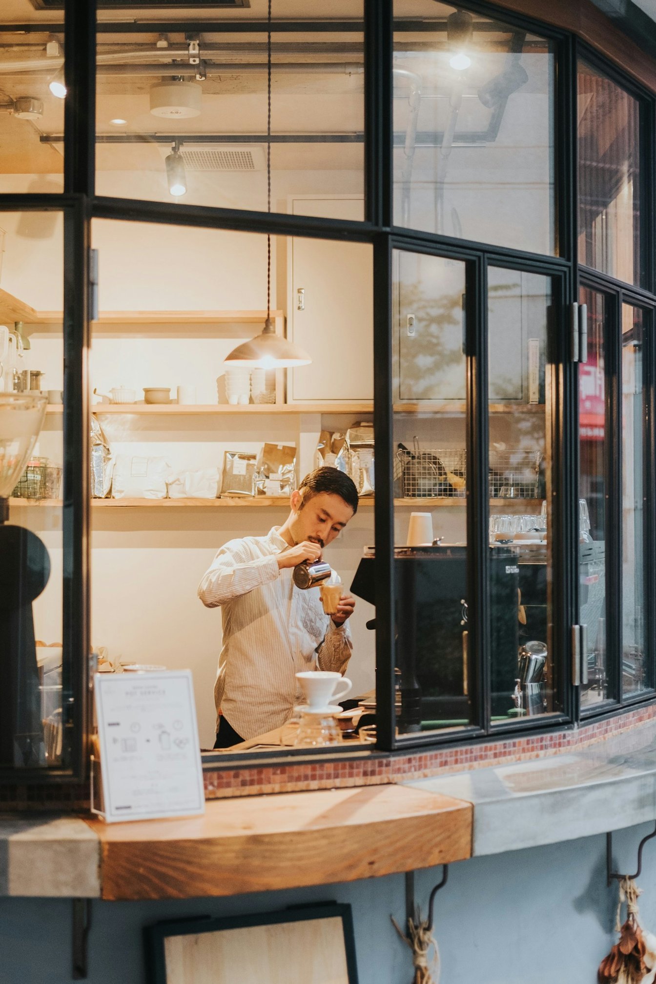 A barista pours steamed milk into a glass inside a café window in warm light