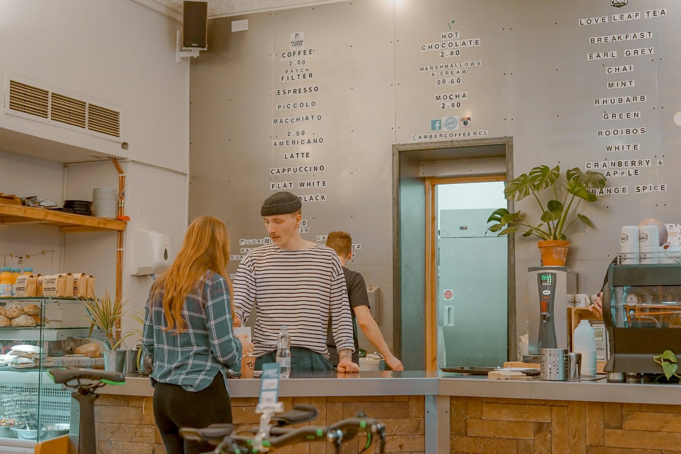 A customer and a barista in a striped top talk across a café counter beneath a handwritten menu